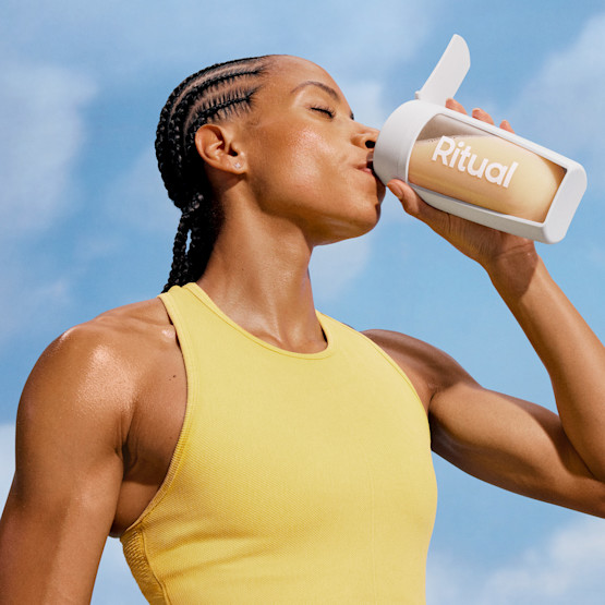 Model in a yellow tank top drinking a protein shaker with Essential Protein Daily Shake. 