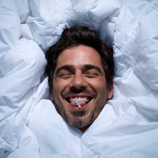 Model surrounded by duvet comforter balancing pill on tongue.